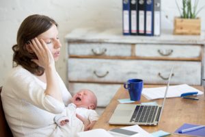 Young businesswoman holding newborn crying babe while sitting with laptop and touching head with pained expression. Career mom suffering from migraine after working on pc and nursing crying child